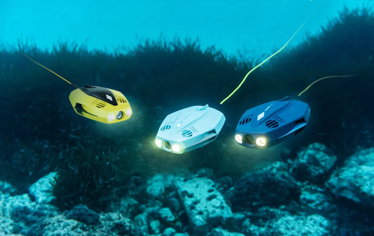 Drei Dory-Drohnen in Gelb, Weiß und Blau mit eingeschalteten Lampen über Seegras und Felsen