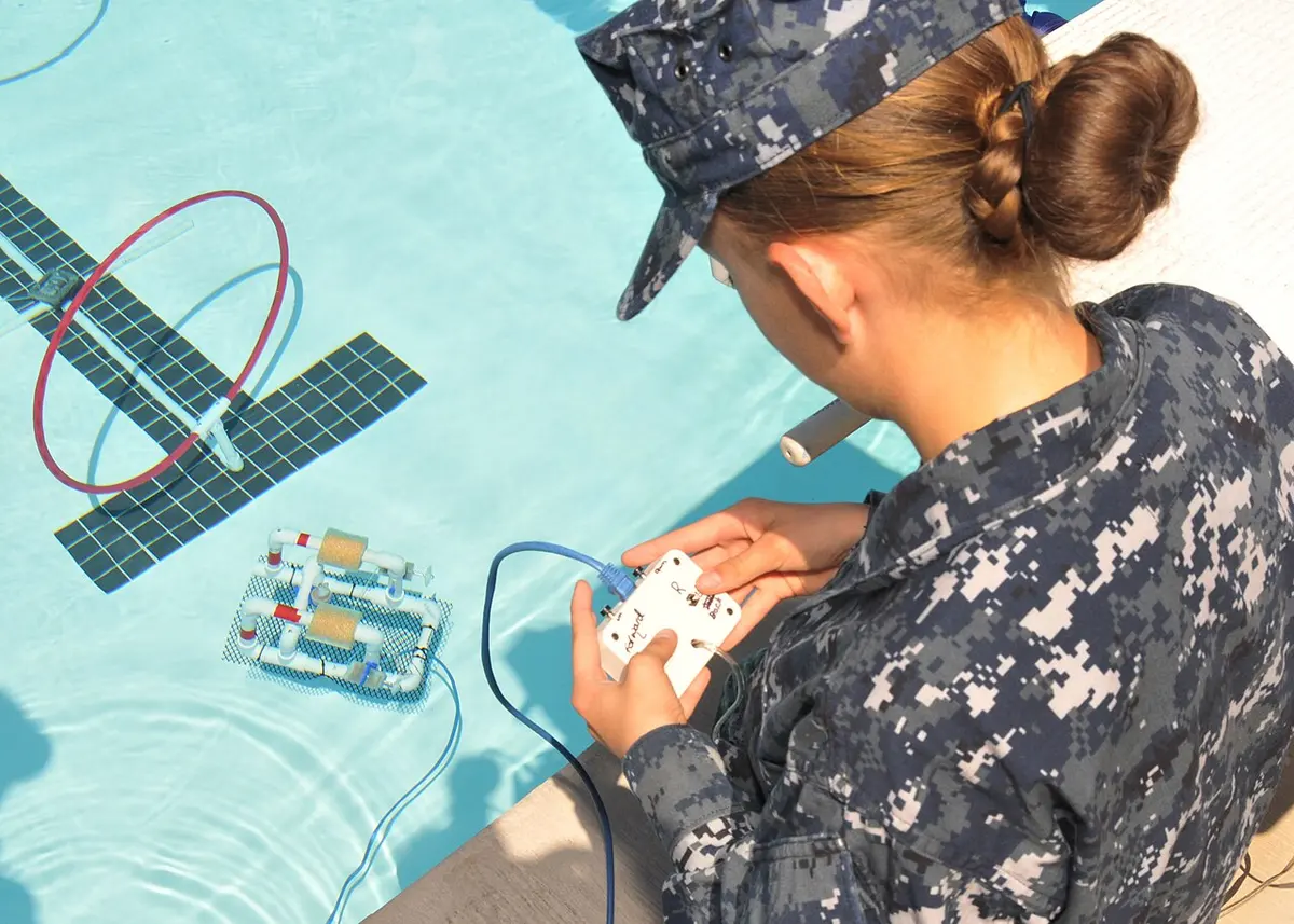 Ein SeaPerch-ROV aus weißen PVC-Rohren mit roten und gelben Schwimmern im Schwimmbecken, eine Person steuert es vom Beckenrand mit einer kleinen Schalterbox